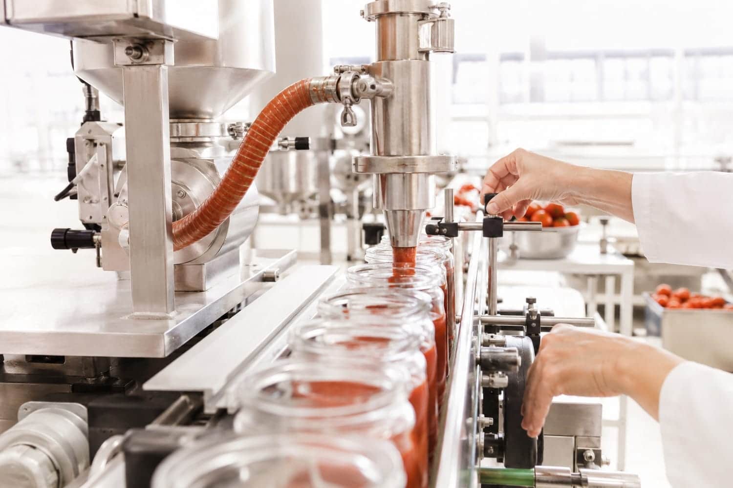 Tomato-red sauce filling line in a bright white production kitchen, hands in frame, daylight from large windows